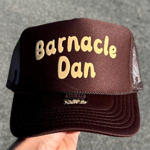 May include: A brown trucker hat featuring a foam front panel with the text Barnacle Dan printed in a light yellow, rounded font. The hat includes a mesh back, a curved brim with decorative stitching, and a small black tag on the lower front.