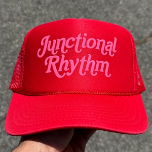 May include: A bright red trucker hat with a pink script that reads "Junctional Rhythm" across the front. The hat features a mesh back and a curved brim. The hat is a vibrant accessory.