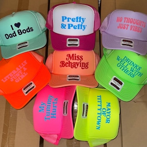 May include: A collection of eight colorful trucker hats featuring various slogans including I Love Dad Bods, Pretty & Petty, No Thoughts Just Vibes, Literally Just A Girl, Miss Behaving, Drinks Well With Others, My Tummy Hurts, and Mayor Of Tittytown.