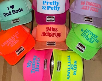 Op de afbeelding: Een collectie van acht kleurrijke trucker-petten met diverse slogans, waaronder I Love Dad Bods, Pretty & Petty, No Thoughts Just Vibes, Literally Just A Girl, Miss Behaving, Drinks Well With Others, My Tummy Hurts en Mayor Of Tittytown.