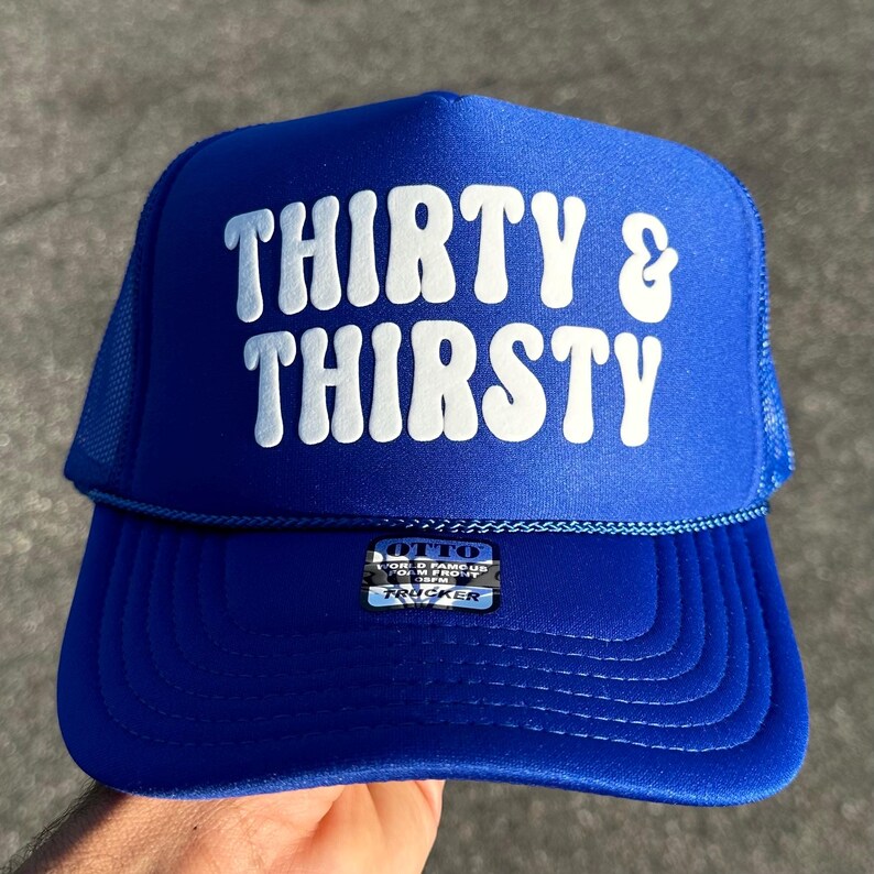 May include: A bright blue trucker hat featuring a foam front panel with the white text THIRTY & THIRSTY in a bold, wavy font. The cap includes a matching blue mesh back, a blue cord detail across the brim, and an Otto brand sticker on the front.
