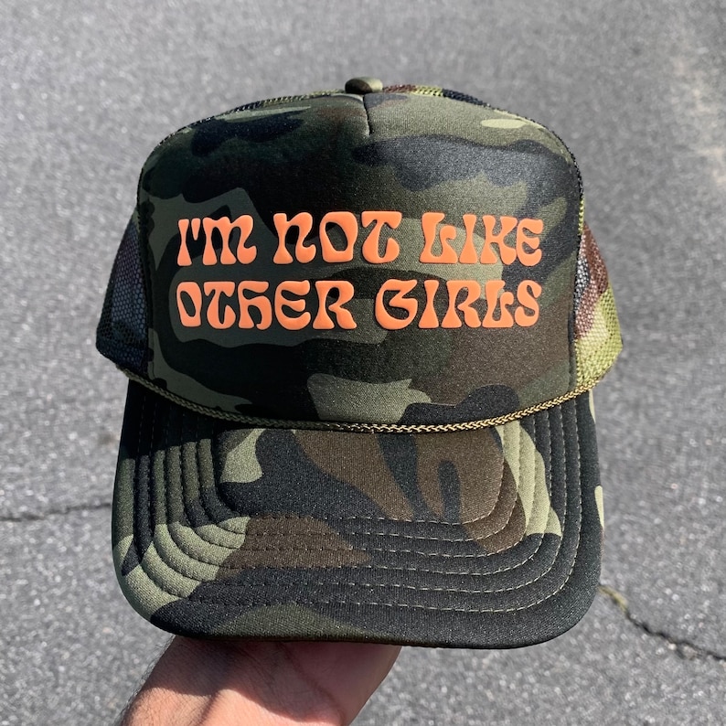 May include: Camouflage trucker hat with the text "I'M NOT LIKE OTHER GIRLS" in orange. The hat features a mesh back and a curved brim. The hat is a mix of green, brown, and black.
