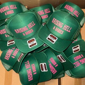 May include: A pile of green trucker hats with pink text that reads "RAISING HELL & BABIES." Each hat has a mesh back and a white tag that says "OTTO" and "FOAM FRONT TRUCKER."