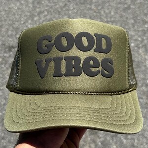 May include: An olive green trucker hat featuring a structured front panel with the text GOOD VIBES printed in bold black lettering. The cap includes a matching olive green brim with decorative stitching and dark mesh side and back panels.