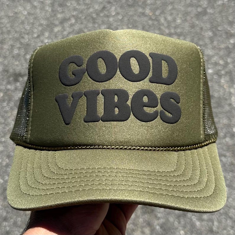 May include: An olive green trucker hat featuring a structured front panel with the text GOOD VIBES printed in bold black lettering. The cap includes a matching olive green brim with decorative stitching and dark mesh side and back panels.