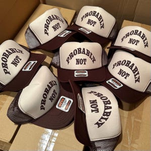 May include: A collection of trucker hats with a brown brim and mesh back. The front of each hat is cream-colored with the words "PROBABLY NOT" printed in brown. The hats are stacked in a cardboard box.