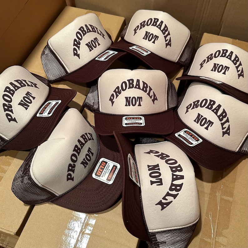 May include: A collection of trucker hats with a brown brim and mesh back. The front of each hat is cream-colored with the words "PROBABLY NOT" printed in brown. The hats are stacked in a cardboard box.