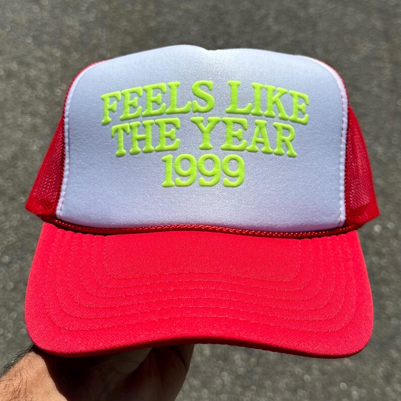 May include: A red and white trucker hat featuring a foam front panel with the neon green text FEELS LIKE THE YEAR 1999 printed in a bold, rounded font. The cap includes a red curved brim and red mesh side and back panels for ventilation.