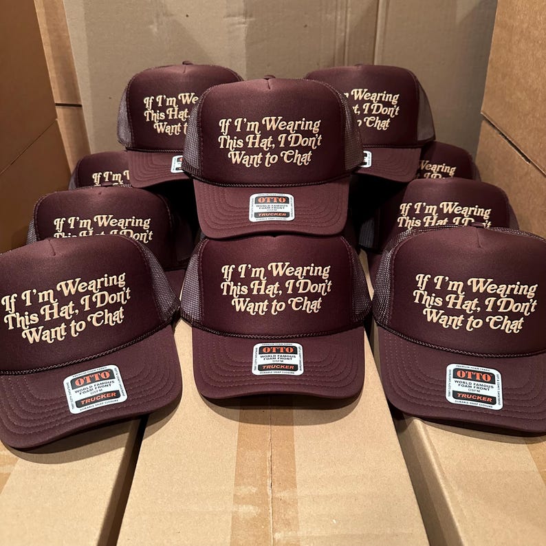 May include: A collection of maroon trucker hats with a mesh back. Each hat features the text "If I'm Wearing This Hat, I Don't Want to Chat" in a white, stylized font. The hats have a curved brim and a tag that reads "OTTO Trucker".