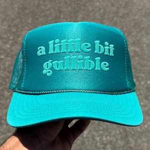 May include: Teal trucker hat with the phrase "a little bit gullible" in raised, matching teal lettering. The hat has a mesh back and a curved brim. A casual accessory.