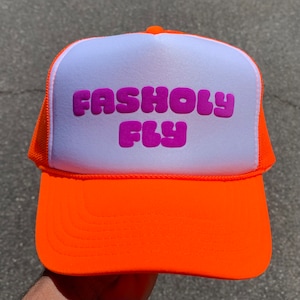 May include: Bright orange trucker hat with a white front panel. The hat features the text "FASHOLY FLY" in a vibrant purple color. The hat has a mesh back and a curved brim.