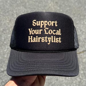 May include: A black trucker hat featuring a foam front panel and a mesh back. The front of the hat displays the text Support Your Local Hairstylist in a tan, serif font. The hat includes a matching black brim with decorative stitching.