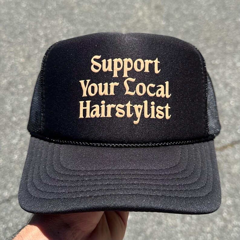 May include: A black trucker hat featuring a foam front panel and a mesh back. The front of the hat displays the text Support Your Local Hairstylist in a tan, serif font. The hat includes a matching black brim with decorative stitching.