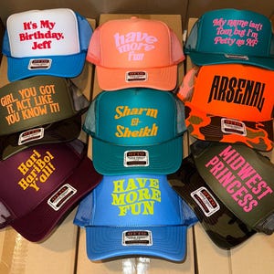 May include: A collection of nine foam front trucker hats in various colors including blue, orange, teal, olive, and maroon. Each hat features unique text prints such as It's My Birthday Jeff, have more fun, Sharm El-Sheikh, and ARSENAL, with mesh backs.
