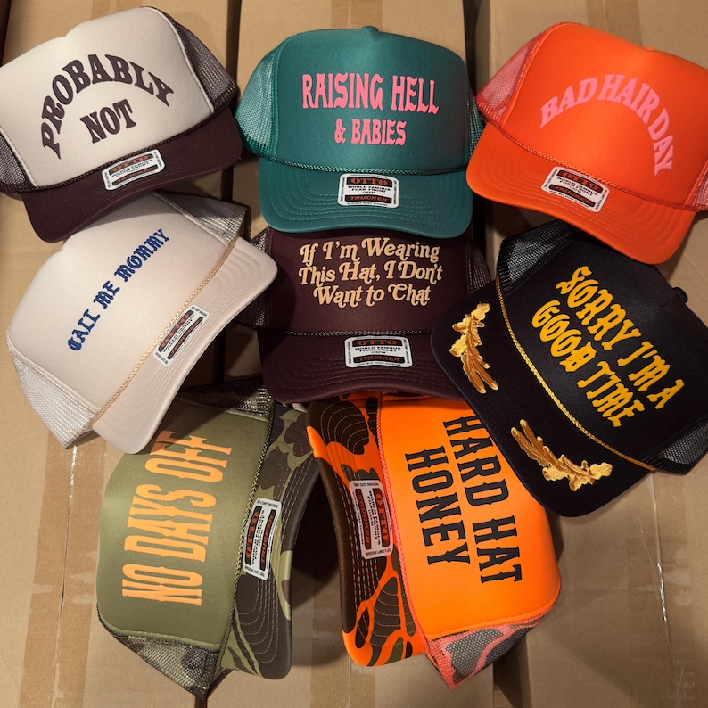 May include: A collection of trucker hats in various colors, including white, orange, green, and black. Each hat features unique text such as "Probably Not," "Raising Hell & Babies," and "Bad Hair Day." The hats have mesh backs and curved brims.