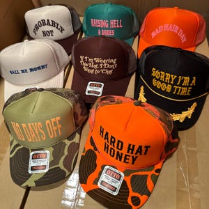 May include: A collection of trucker hats in various colors, including black, orange, and green. Each hat features unique text such as "Probably Not," "Raising Hell & Babies," and "Hard Hat Honey." The hats have mesh backs and foam fronts.