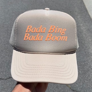 May include: Tan trucker hat with a mesh back and a curved brim. The hat features the orange text "Bada Bing Bada Boom" on the front panel. The hat is a neutral color and suitable for various casual outfits.