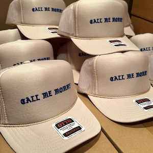 May include: A collection of beige trucker hats with the phrase "CALL ME MOMMY" embroidered in blue. Each hat features a mesh back, a curved brim, and a rope detail. The hats have a "OTTO" label.