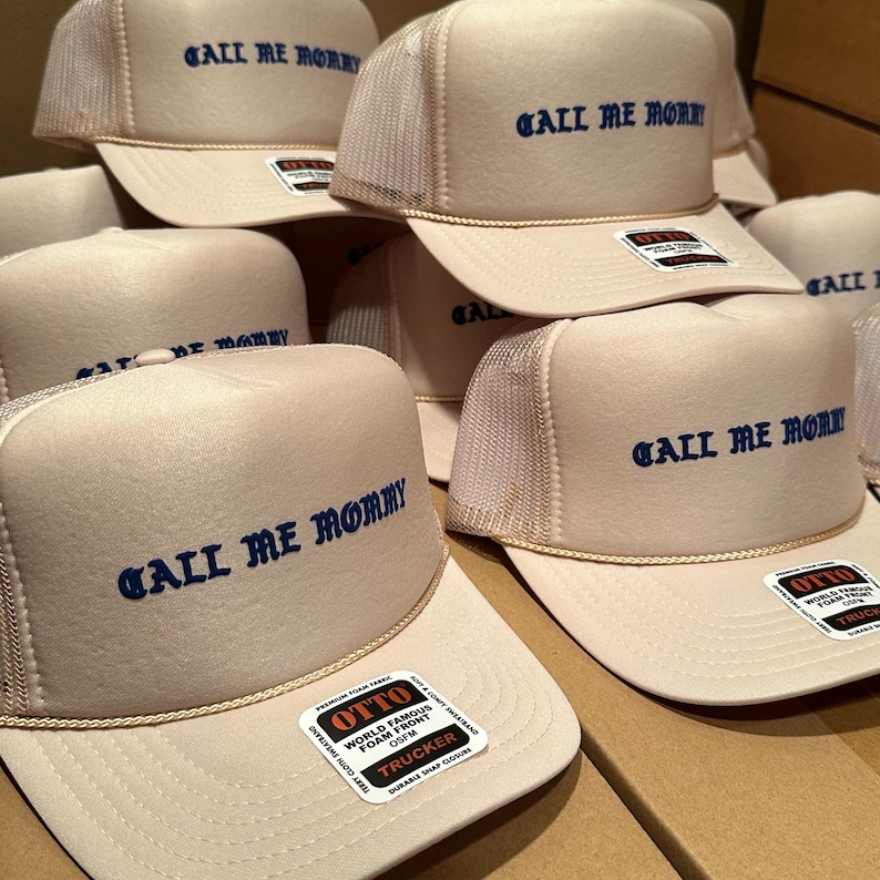 May include: A collection of beige trucker hats with the phrase "CALL ME MOMMY" embroidered in blue. Each hat features a mesh back, a curved brim, and a rope detail. The hats have a "OTTO" label.