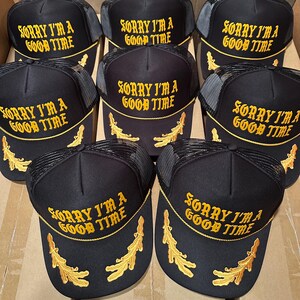 May include: Black trucker hats with the text "SORRY I'M A GOOD TIME" embroidered in gold. The hats have a mesh back and a curved brim with gold leaf accents. A collection of hats.