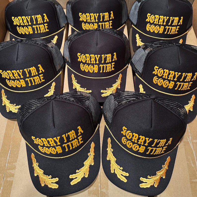 May include: Black trucker hats with the text "SORRY I'M A GOOD TIME" embroidered in gold. The hats have a mesh back and a curved brim with gold leaf accents. A collection of hats.