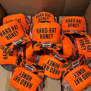 May include: A pile of orange trucker hats with the text "HARD HAT HONEY" in black block letters. The hats have a camouflage pattern on the brim and a mesh back. The hats are stacked in a cardboard box.