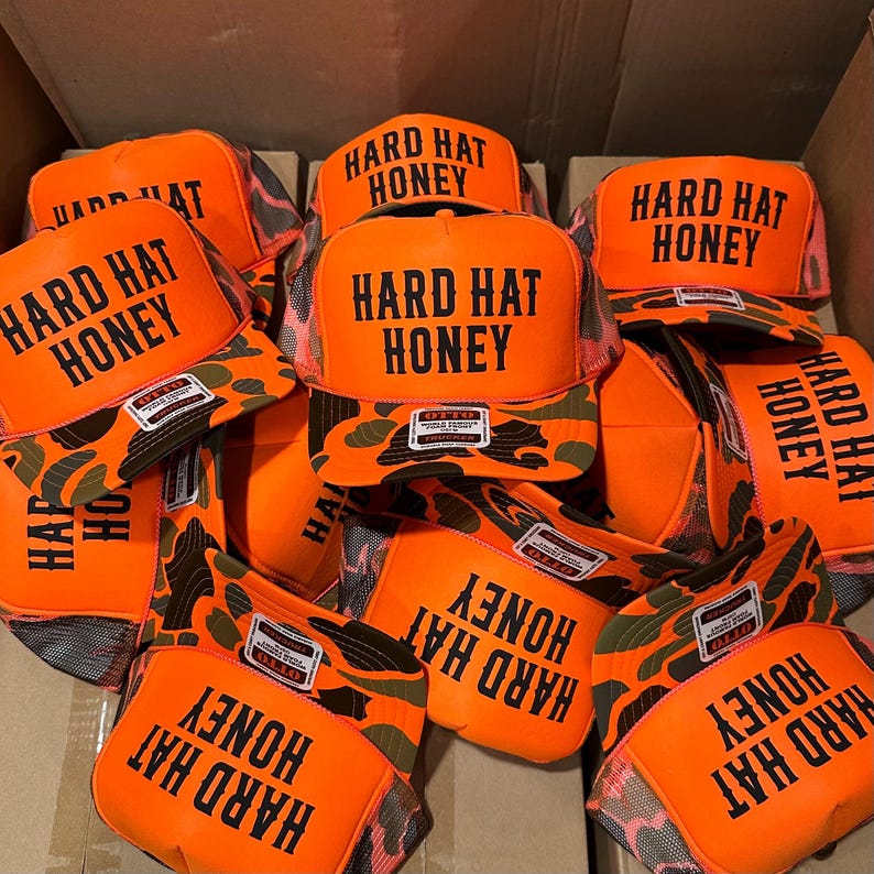 May include: A pile of orange trucker hats with the text "HARD HAT HONEY" in black block letters. The hats have a camouflage pattern on the brim and a mesh back. The hats are stacked in a cardboard box.