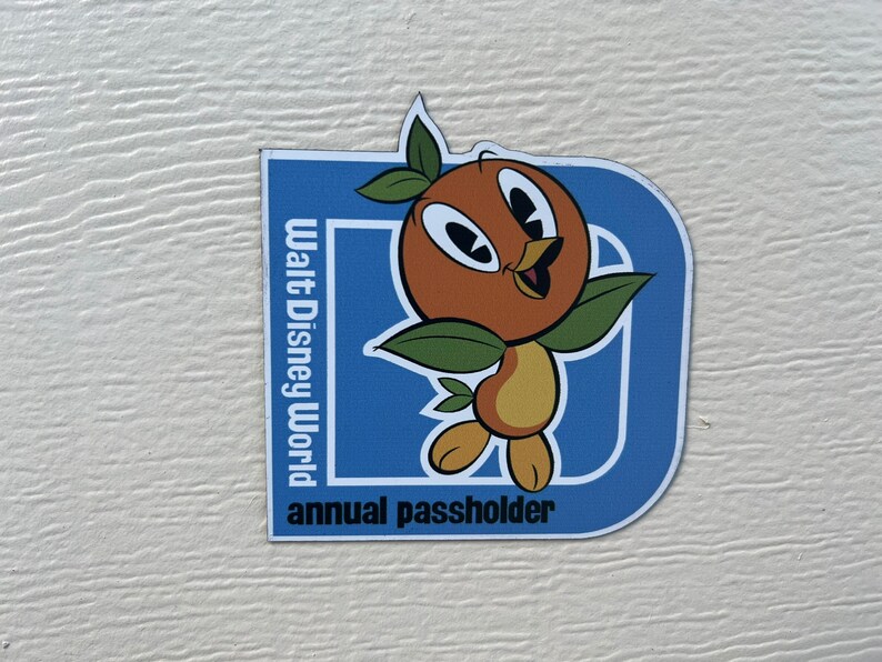 Pu&ograve; includere: Un magnete blu e bianco con un uccello arancione dei cartoni animati e il testo "Walt Disney World annual passholder".