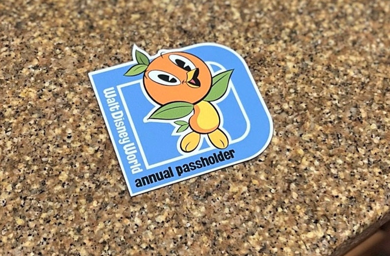 Pu&ograve; includere: Adesivo blu e arancione con un personaggio di cartone animato di un uccello arancione. L'adesivo dice "Walt Disney World annual passholder".