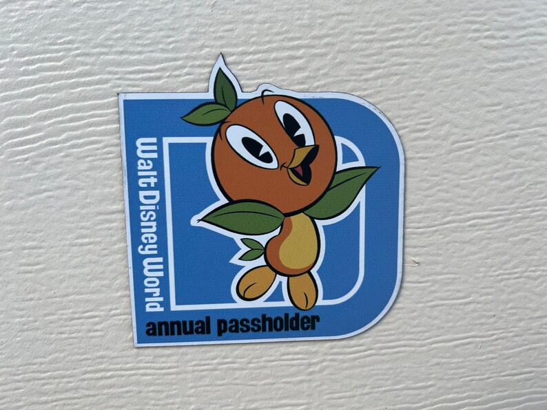 Pu&ograve; includere: Un magnete blu e bianco con il logo di Walt Disney World e le parole "annual passholder". Un uccello arancione dei cartoni animati con foglie verdi &egrave; al centro del magnete.