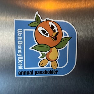 Pu&ograve; includere: Un magnete per pass annuale Walt Disney World blu e bianco con un uccello arancione dei cartoni animati con foglie verdi.