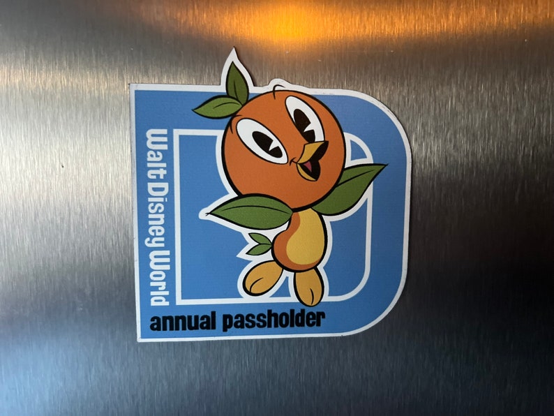 Pu&ograve; includere: Un magnete per pass annuale Walt Disney World blu e bianco con un uccello arancione dei cartoni animati con foglie verdi.