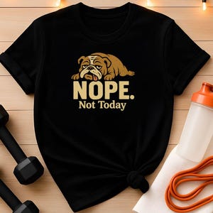 Könnte beinhalten: Schwarzes T-Shirt mit einem braunen Bulldoggen-Motiv und dem Text "NOPE. Not Today" in Gold. Das T-Shirt ist unten geknotet. Eine Wasserflasche, ein Handtuch und ein Widerstandsband sind im Hintergrund. Fitness-Thema.