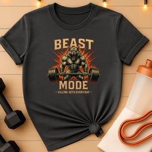 Puede incluir: Camiseta gris oscuro con un gráfico de un lobo musculoso levantando una barra, las palabras "BEAST MODE" y "KILLING SETS EVERYDAY". Incluye pesas, una botella de agua y una banda de resistencia naranja.