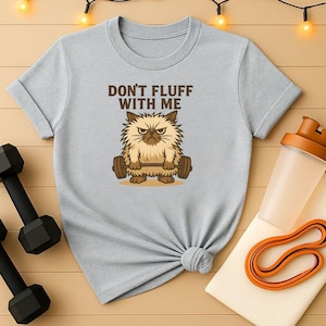 Könnte beinhalten: Hellgraues T-Shirt mit dem Text "DON'T FLUFF WITH ME" und einer Cartoon-Katze, die Gewichte hebt. Die Katze ist braun und hat einen wütenden Gesichtsausdruck. Eine schwarze Hantel, eine Wasserflasche und ein orangefarbenes Widerstandsband sind ebenfalls zu sehen.