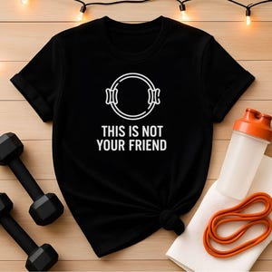 Könnte beinhalten: Schwarzes T-Shirt mit dem weißen Text "THIS IS NOT YOUR FRIEND" und einem weißen Grafikdesign. Auf dem Bild sind Fitness-Zubehör wie Kurzhanteln, eine Wasserflasche und Widerstandsbänder zu sehen.