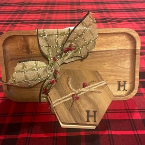 Puede incluir: Una bandeja de servir de madera con un lazo decorativo y un pequeño posavasos de madera con la letra "H" grabada. La bandeja y el posavasos están sobre una superficie a cuadros rojos y negros.