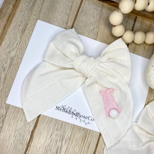 May include: White linen bow with a pink embroidered bunny and a white pom-pom tail. The bow is on a wooden surface with the text "StellaAndRoseCo" printed on a white piece of paper.