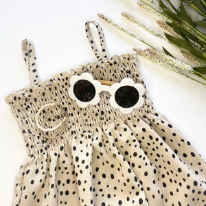 May include: A beige and black polka dot dress with thin straps. The dress has a smocked bodice and is made of a lightweight fabric. A pair of white flower-shaped sunglasses with black lenses is placed on top of the dress.
