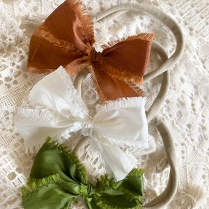 May include: Three hair bows with frayed edges. The bows are brown, white, and green. The bows are tied around elastic headbands.