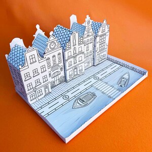 May include: A paper model of a row of houses with blue roofs and white walls. The houses are arranged in a row on a white base with a black and white design of a street and canal. Two small boats are on the canal.