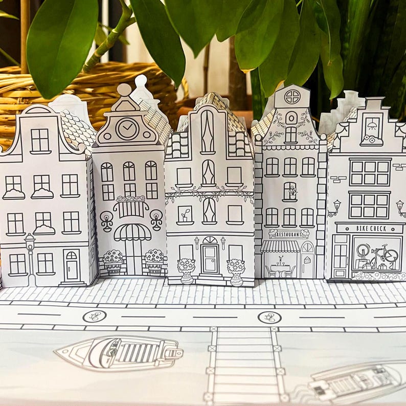 May include: Black and white printable papercraft model of a row of five European-style houses with a boat and a ladder in the foreground. The houses have details like windows, doors, and signs that say "Restaurant" and "Bike Check".