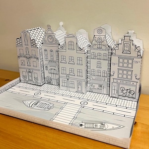 May include: A black and white paper model of a European town with buildings, a canal, and boats. The buildings have details like windows, doors, and roofs. The canal has a bridge and a boat on it. The model is made of paper and is designed to be assembled.