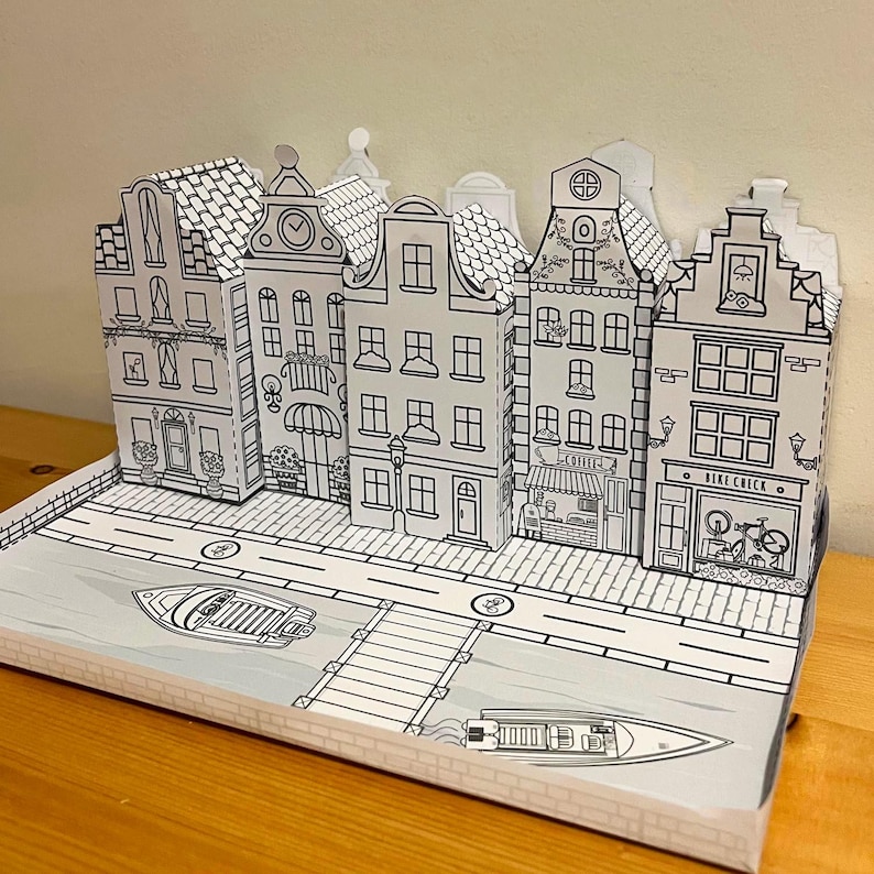May include: A black and white paper model of a European town with buildings, a canal, and boats. The buildings have details like windows, doors, and roofs. The canal has a bridge and a boat on it. The model is made of paper and is designed to be assembled.