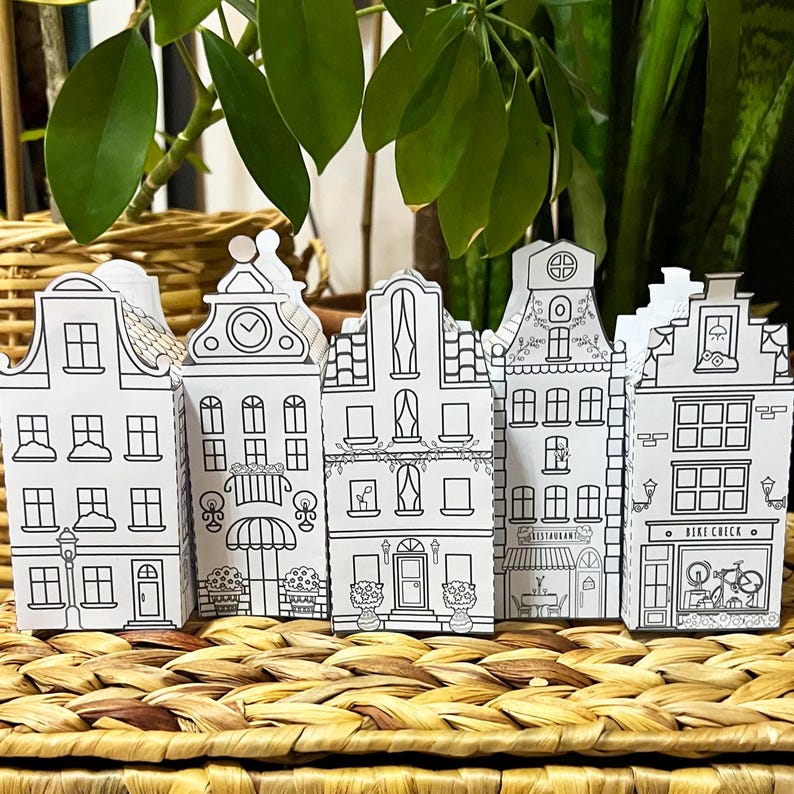 May include: Five black and white paper model houses with different details, including windows, doors, and a clock. The houses are arranged in a row on a woven wicker surface.