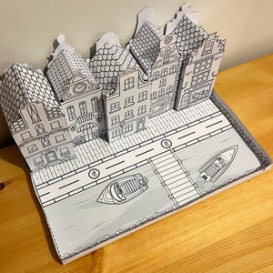 May include: A paper model of a Dutch village with houses, a canal, and boats. The model is made of white paper and is unpainted.