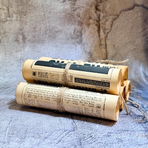 May include: A stack of three rolled parchment scrolls tied together with natural jute twine. The scrolls feature printed black text and graphics resembling a vintage newspaper, with visible columns and headlines on the aged, off-white paper.