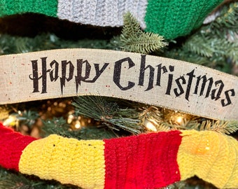 Könnte beinhalten: Ein beiges Band mit dem schwarzen Schriftzug Happy Christmas in einer stilisierten Schriftart, platziert zwischen zwei gehäkelten Schals. Der obere Schal hat grüne und graue Streifen, der untere Schal hat rote und gelbe Streifen.