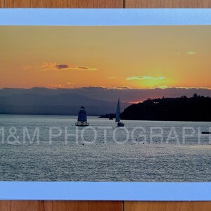 May include: A scenic sunset over a body of water with a lighthouse and sailboat in the distance. The sky is a vibrant orange and yellow, with clouds reflecting the warm light. The water is calm and still, reflecting the colours of the sky.