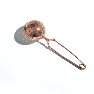 May include: A rose gold tea infuser with a round mesh ball and a long handle. The handle has a spring-loaded clip to secure it to the side of a mug or teapot.